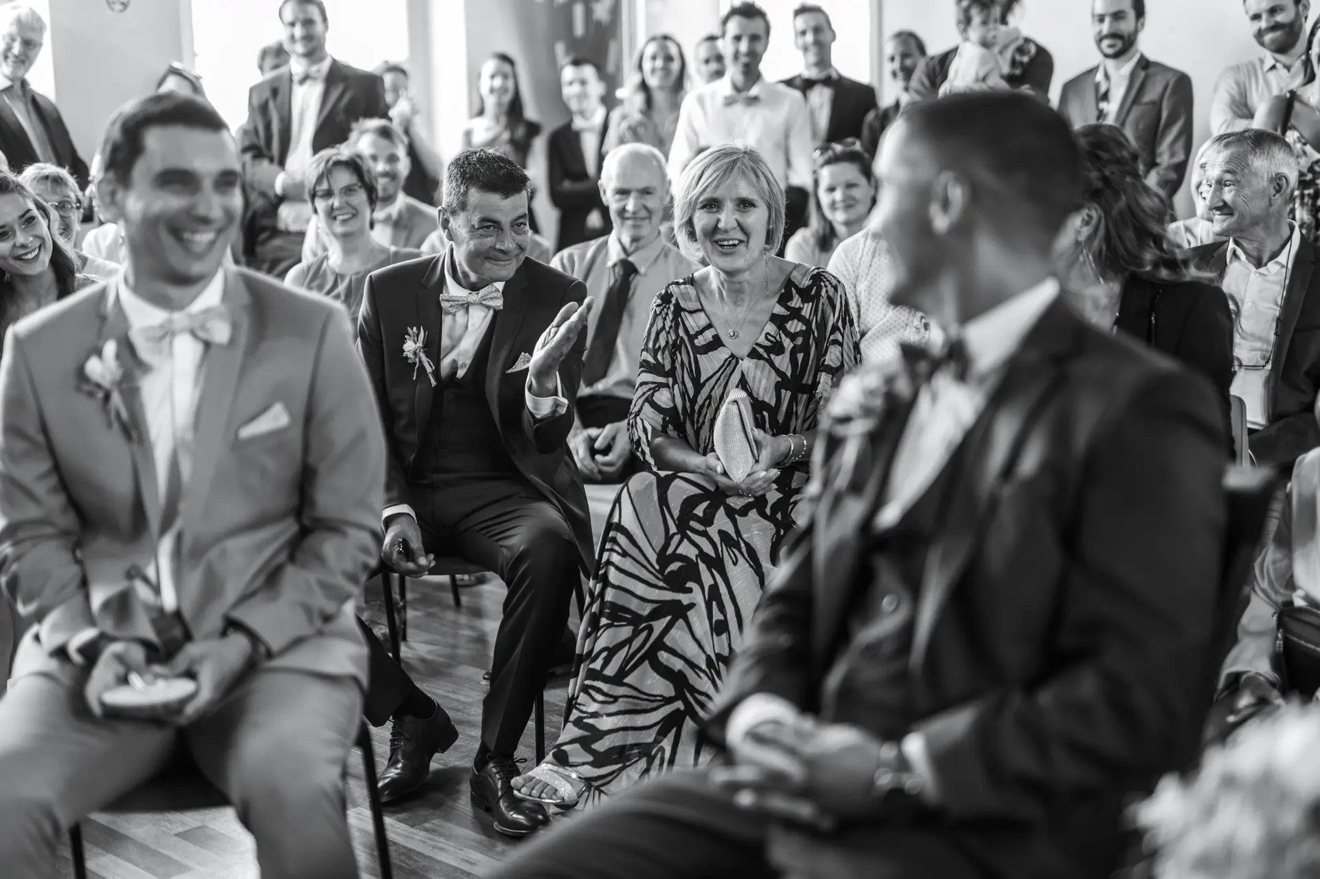 Reportage de mariage noir et blanc à Bonnelles : Focus sur les parents et invités au deuxième rang qui rient aux éclats en regardant le marié flou au premier plan durant la cérémonie civile.