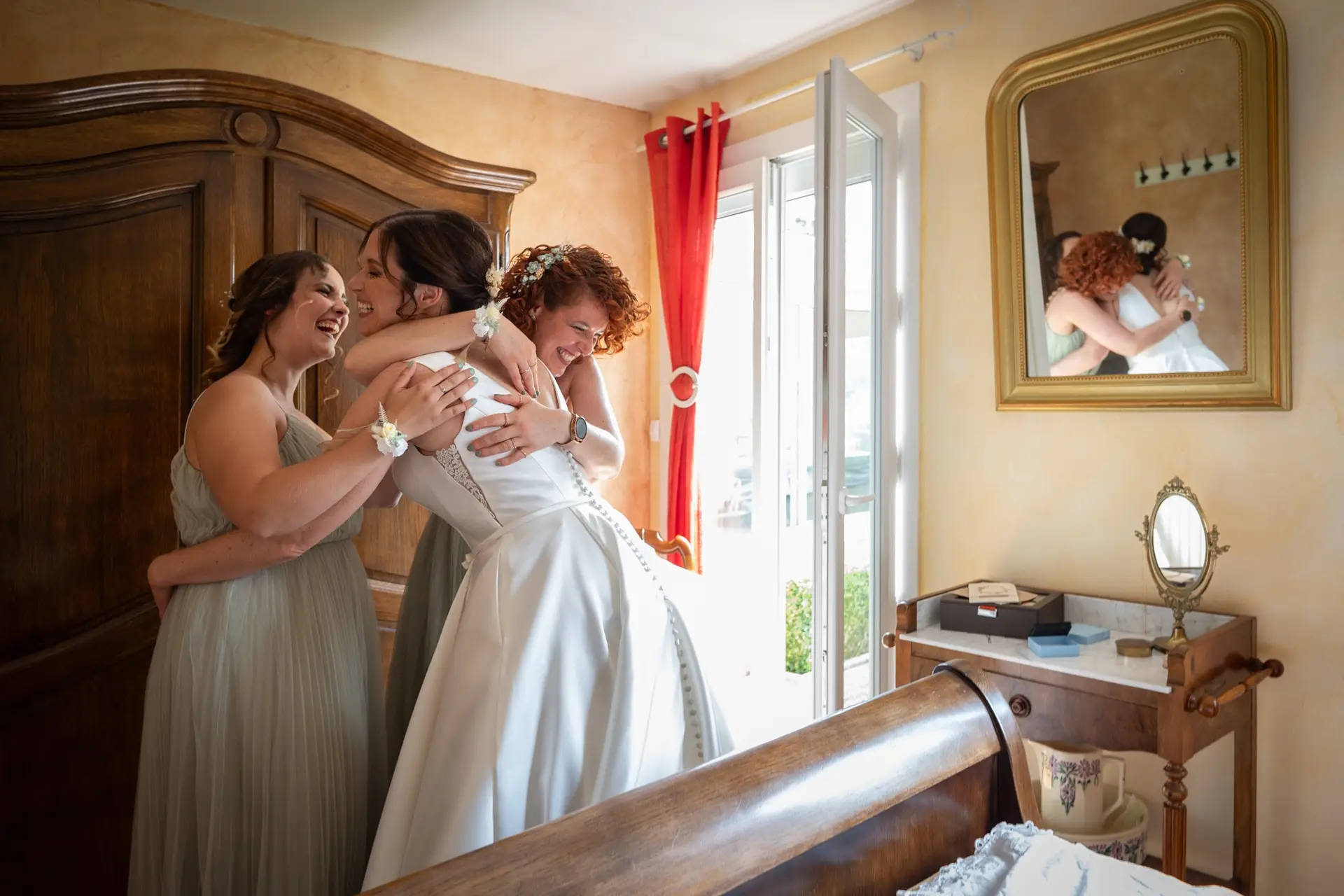 Photographe mariage Eure 27 : étreinte joyeuse entre la mariée et ses demoiselles d'honneur dans une chambre lumineuse lors des préparatifs.