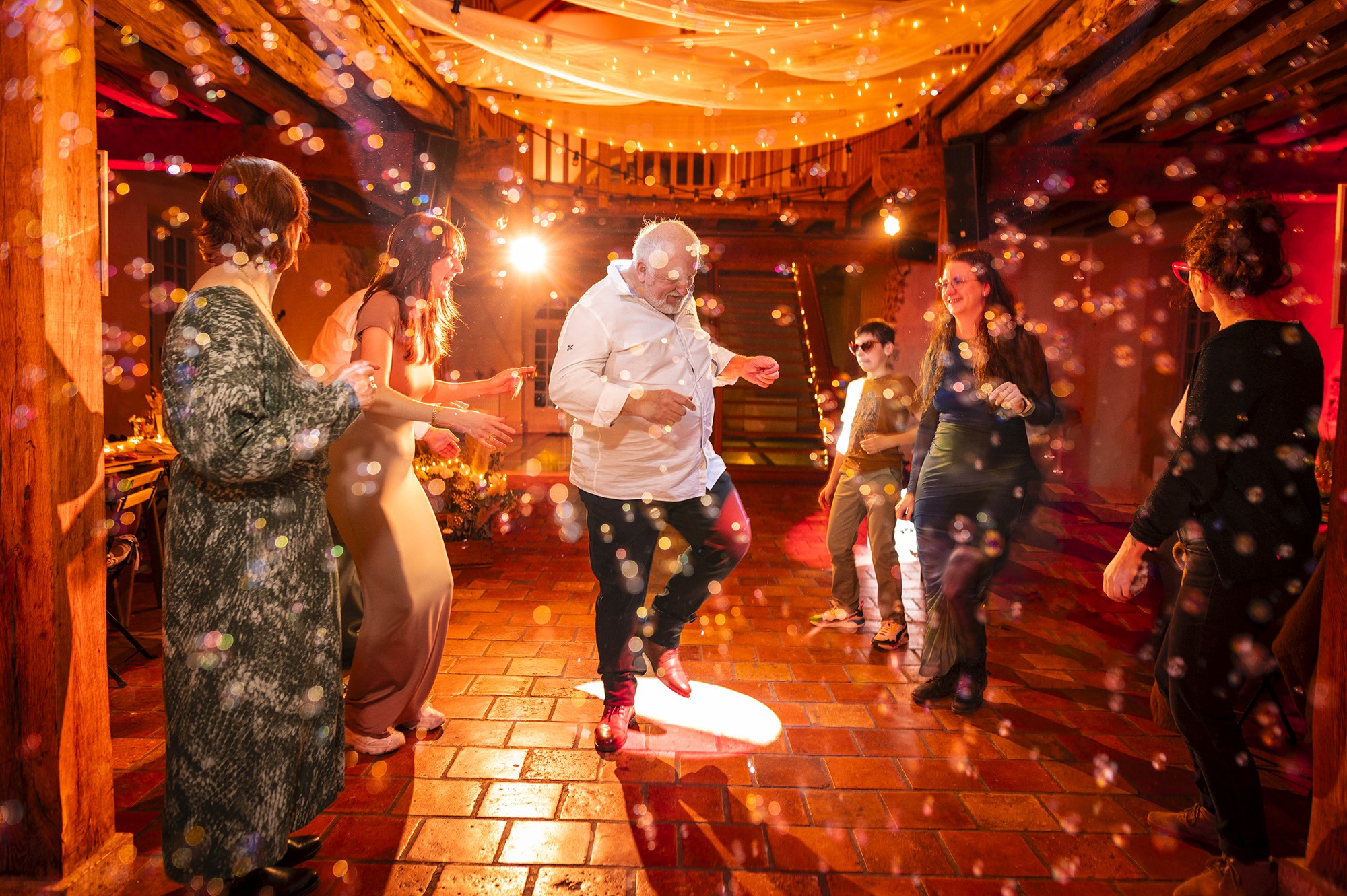 Photographe mariage Moulin de Lambouray - Invités dansant sous une pluie de bulles et des lumières ambrées dans la salle de réception.