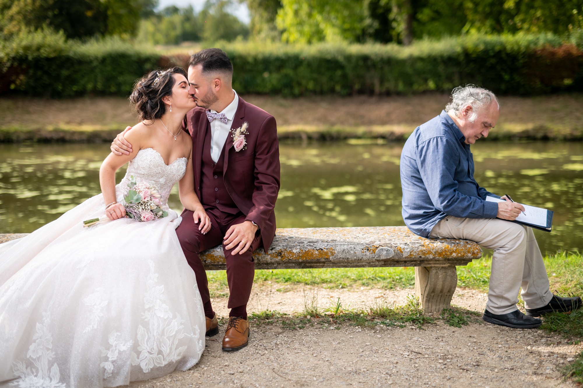 Photographe mariage Rambouillet - Couple de mariés s'embrassant sur un banc en pierre à côté d'un homme qui écrit, dans le parc du Château.